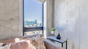 Une chambre moderne dotée d’une grande fenêtre, d’un canapé agrémenté de coussins à motifs, d’une petite table sur laquelle sont posés une plante et une sculpture, et qui offre une vue sur un paysage urbain avec des montagnes à l’horizon.