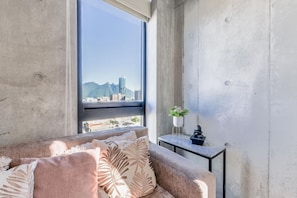 Ein modernes Zimmer mit einem großen Fenster, einem Sofa mit gemusterten Kissen, einem kleinen Tisch mit Pflanze und Skulptur sowie Blick auf eine Stadtlandschaft mit Bergen.