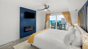 Una habitación de hotel moderna con una cama grande, un televisor de pantalla plana montado en una pared azul, un ventilador de techo y un balcón con vistas a la ciudad.
