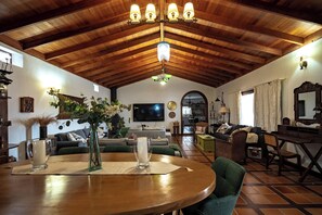Een ruime woonkamer met een houten plafond, een eethoek met een ronde tafel, en een aan de muur bevestigde televisie.
