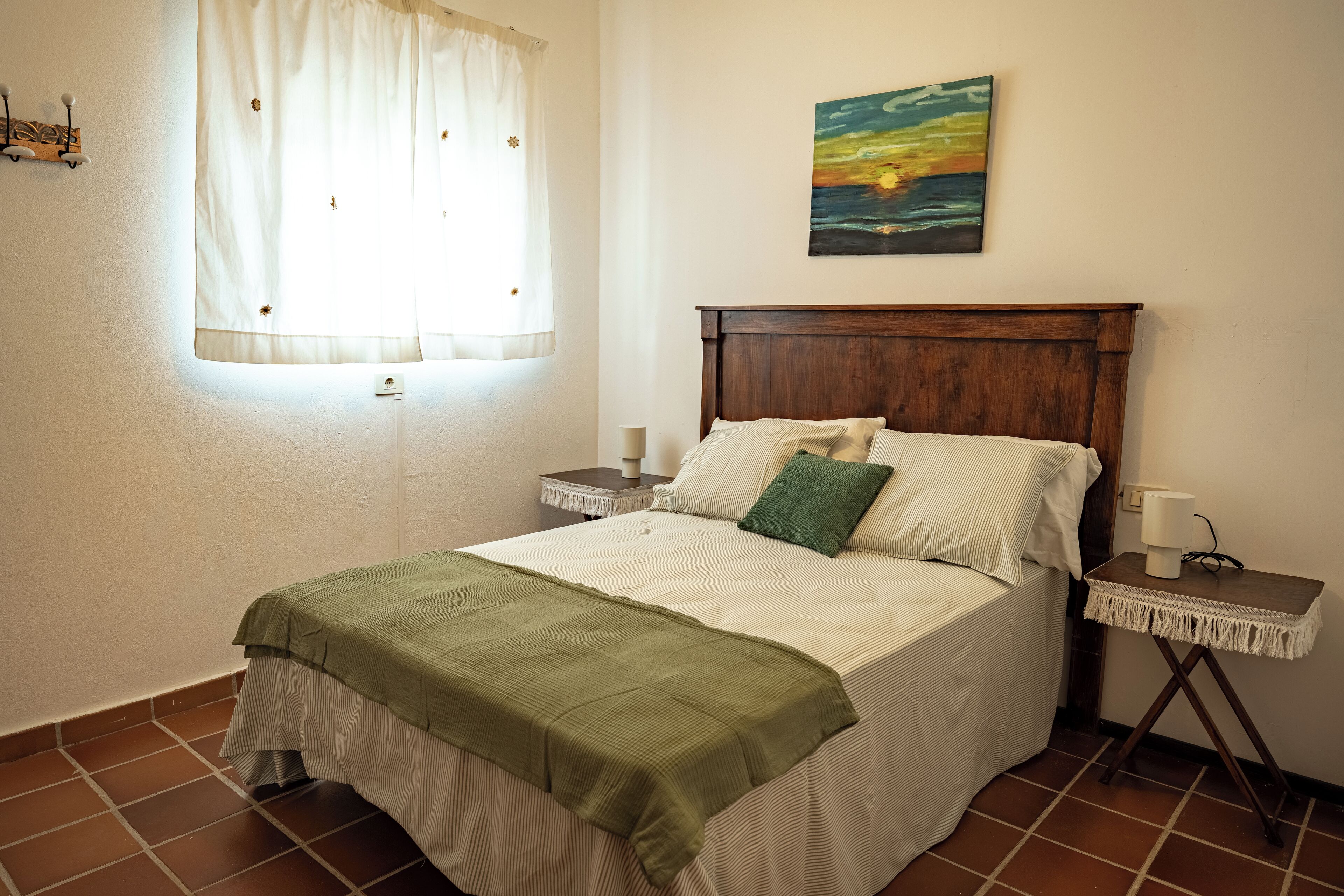 Une chambre à coucher avec un lit, une table de chevet, une lampe, une fenêtre avec des rideaux et un tableau au mur.