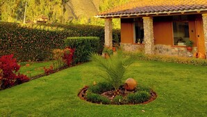 A house with a stone foundation and a tiled roof, surrounded by a well-maintained garden with a palm tree and flower beds.