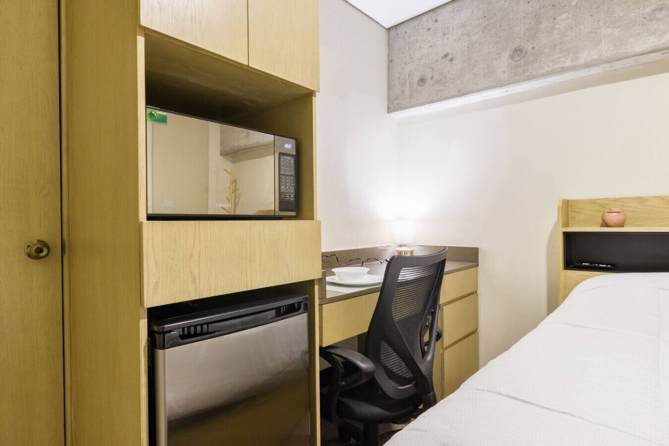 Een compacte hotelkamer met een bed, een bureau met stoel, een magnetron en een klein kastje.