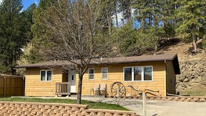 A single-story wooden cabin with a front porch, a small attached garage, and a tree in the front yard. - Rocky Cliff 3 bedroom/2 bath home located in the town of Hill City! (Hill City)