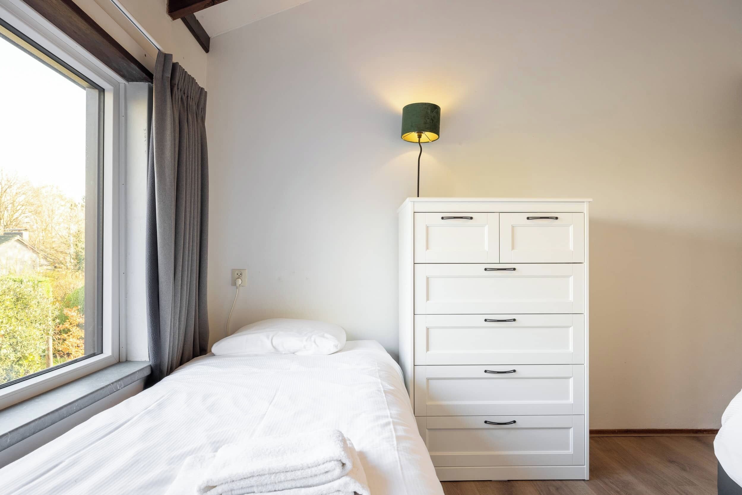 Een slaapkamer met een bed, een witte dressoir, een wandlamp en een raam met gordijnen.