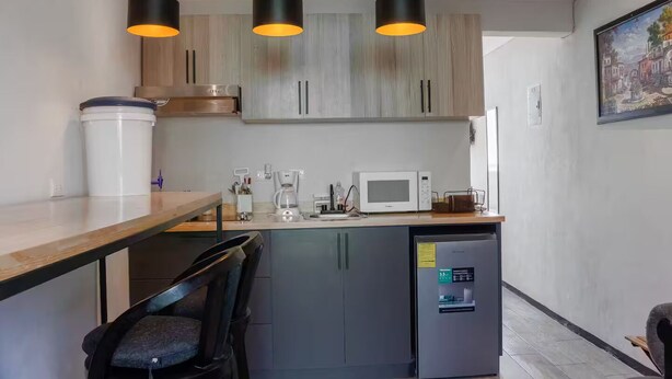 A compact kitchen with a microwave, coffee maker, and a water dispenser. There are three pendant lights, a wooden table, and a chair.