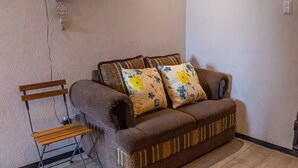 A brown sofa with floral cushions, a wooden chair, and a wall with a crack and decorative plates.