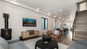 A modern living room with a fireplace, a flat-screen TV, and a dining area with a staircase. - Private Hot Tub | Steps to Trails & Town | Foosball (Winter Park)