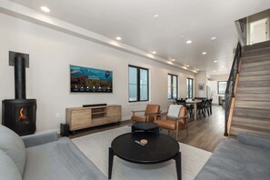 A modern living room with a fireplace, a flat-screen TV, and a dining area with a staircase. - Private Hot Tub | Steps to Trails & Town | Foosball (Winter Park)