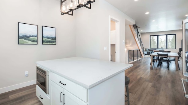 A modern kitchen with white cabinetry, a built-in oven, and a central island. There are two framed landscape paintings on the wall, a staircase leading to a loft area, and a dining area with a large window. - Private Hot Tub | Steps to Trails & Town | Foosball (Winter Park)