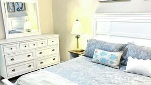Une chambre à coucher comprenant un lit, une commode, une table de chevet, une lampe et deux œuvres d’art murales.