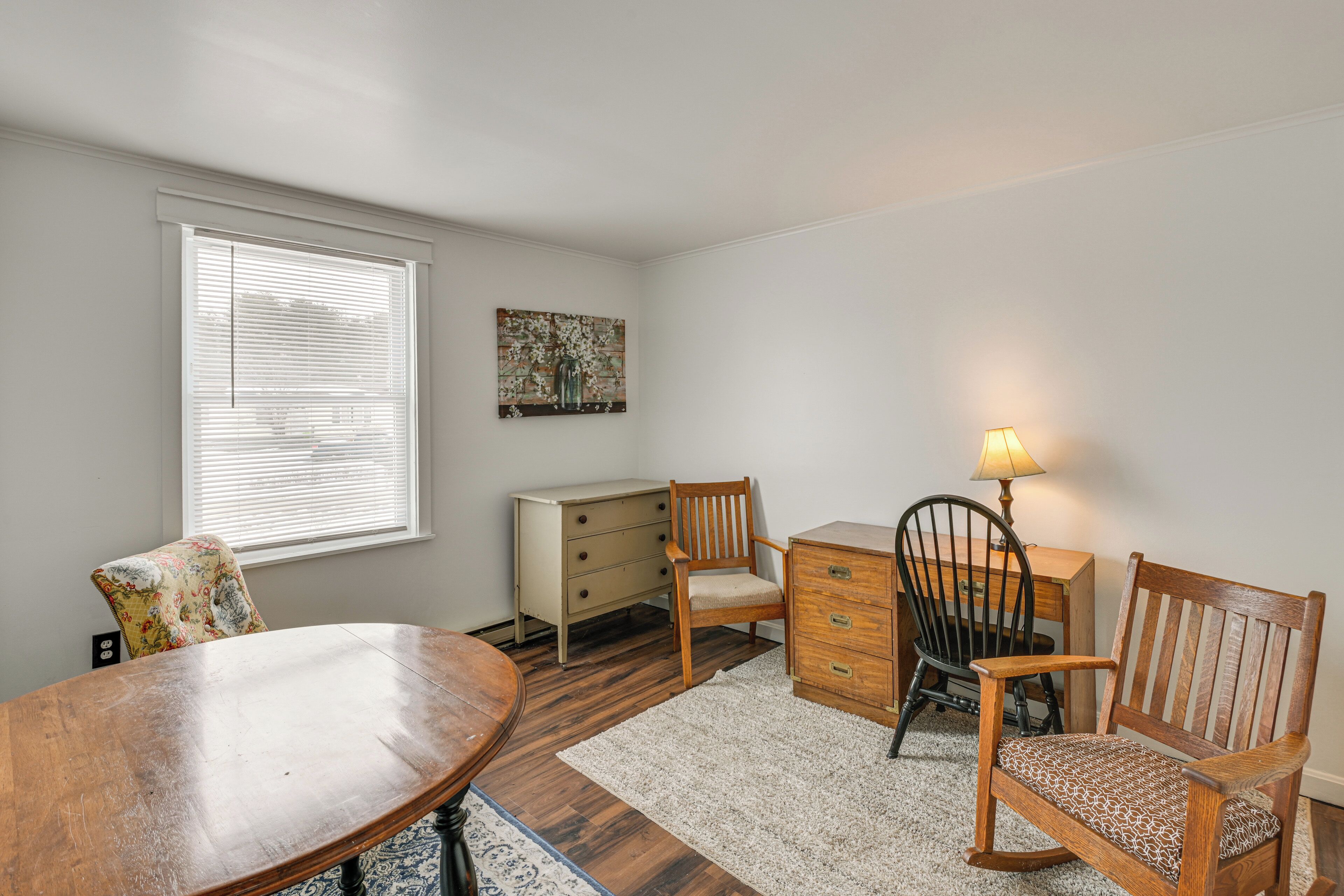 A room with a wooden dining table, two wooden chairs, a small wooden desk, a lamp, and a white dresser.