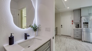 A modern bathroom with a large oval mirror, a white sink, and a potted plant. - Cozy Coachella Valley Home! Golf, Shop + Dine (Indio)