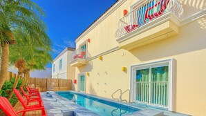 A poolside area with a pool, red chairs, and a building with a balcony. - Steps to the Beach Great Value Pool Home (South Padre Island)