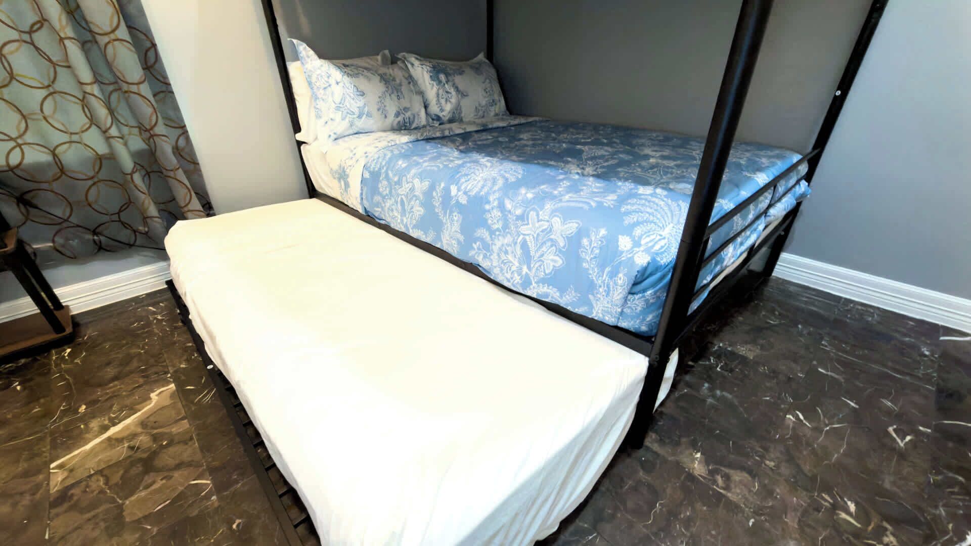 A bunk bed with a blue patterned bedspread and white pillows, a white bench, and a dark marble floor.