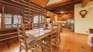 Une table à manger en bois avec des chaises, une cuisine avec des meubles en bois, une horloge au mur et une fenêtre avec des stores.