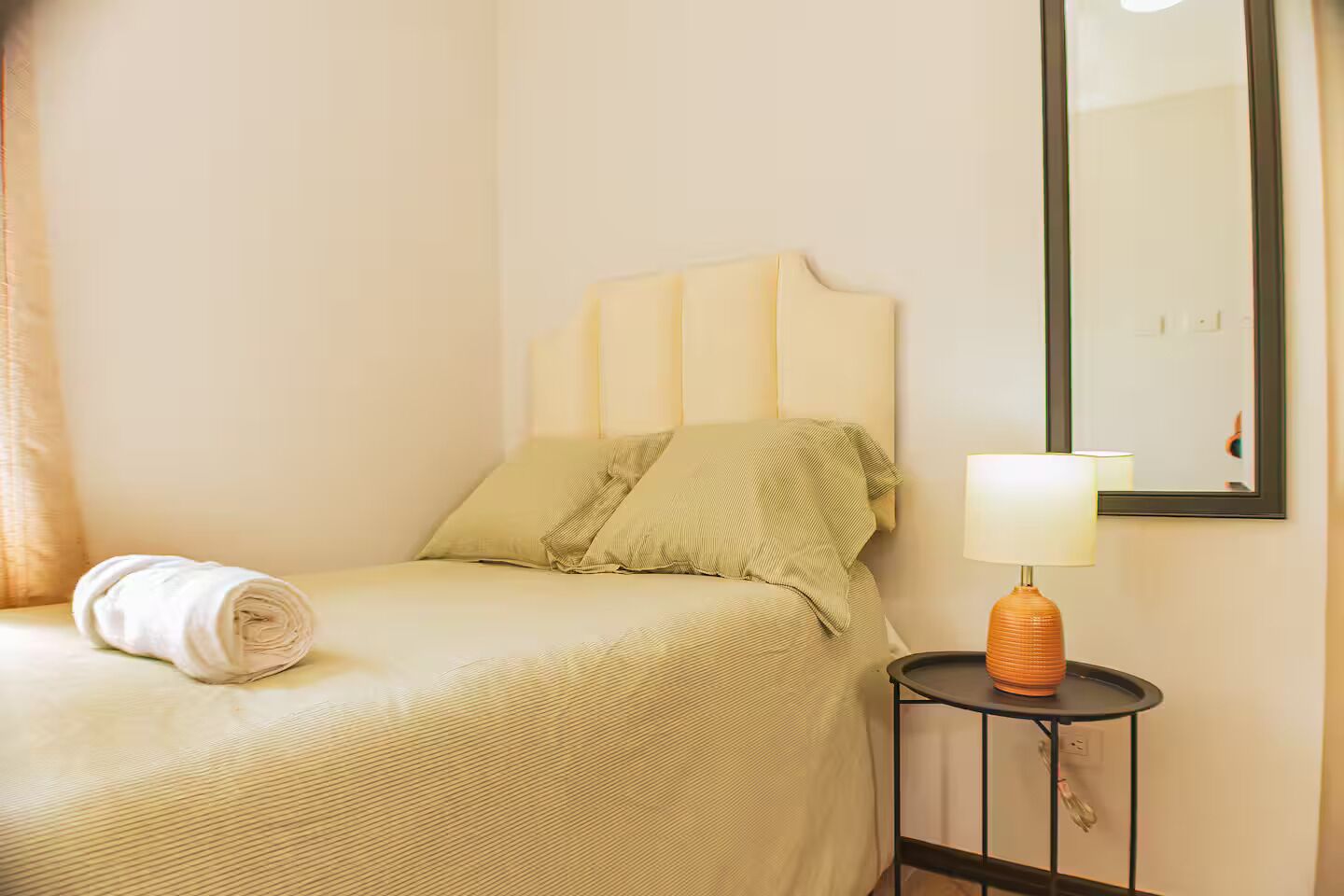 Un lit bien fait, avec une serviette pliée, une lampe de chevet et un miroir accroché au mur.