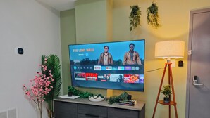 Ein Wohnzimmer mit einem Flachbildfernseher, der 'The Long Walk' auf Netflix zeigt, einer dunklen Holzkommode, einer Stehlampe und Zierpflanzen.