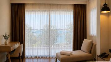 A hotel room with a bed, a desk, a chair, a bedside table, a lamp, and a view of a body of water.