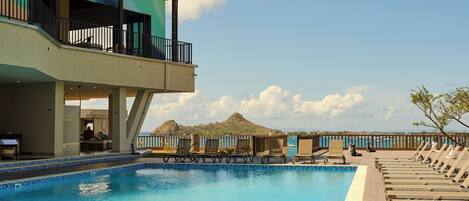 A swimming pool with lounge chairs, a building with balconies, and a clear sky.