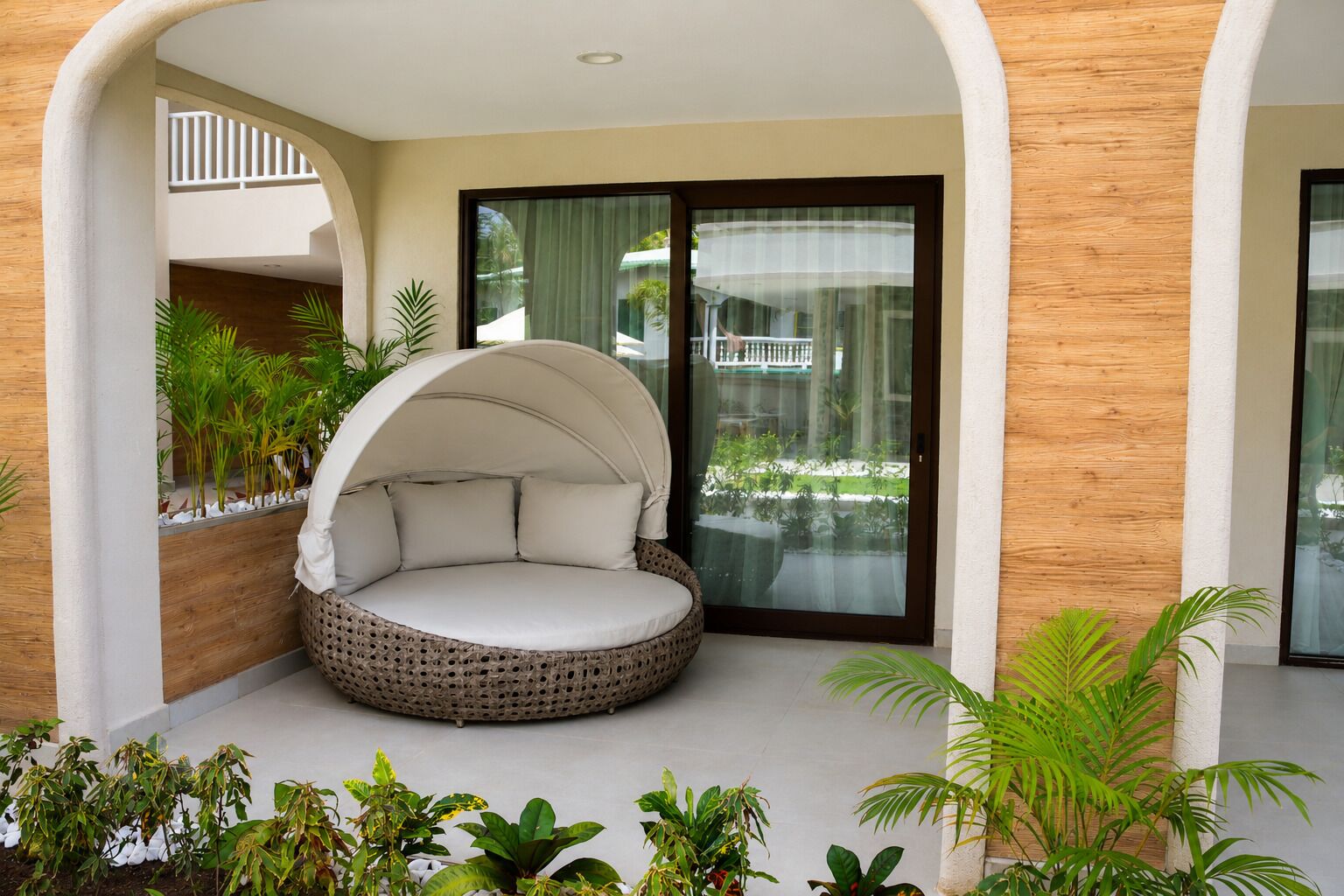 A round, cushioned seating area with a canopy, surrounded by plants and a view of a building through a glass door.