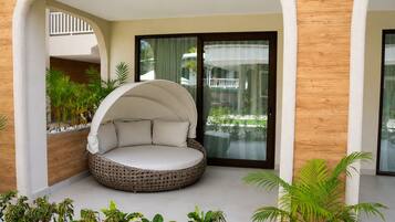 A round, cushioned seating area with a canopy, surrounded by plants and a view of a building through a glass door.