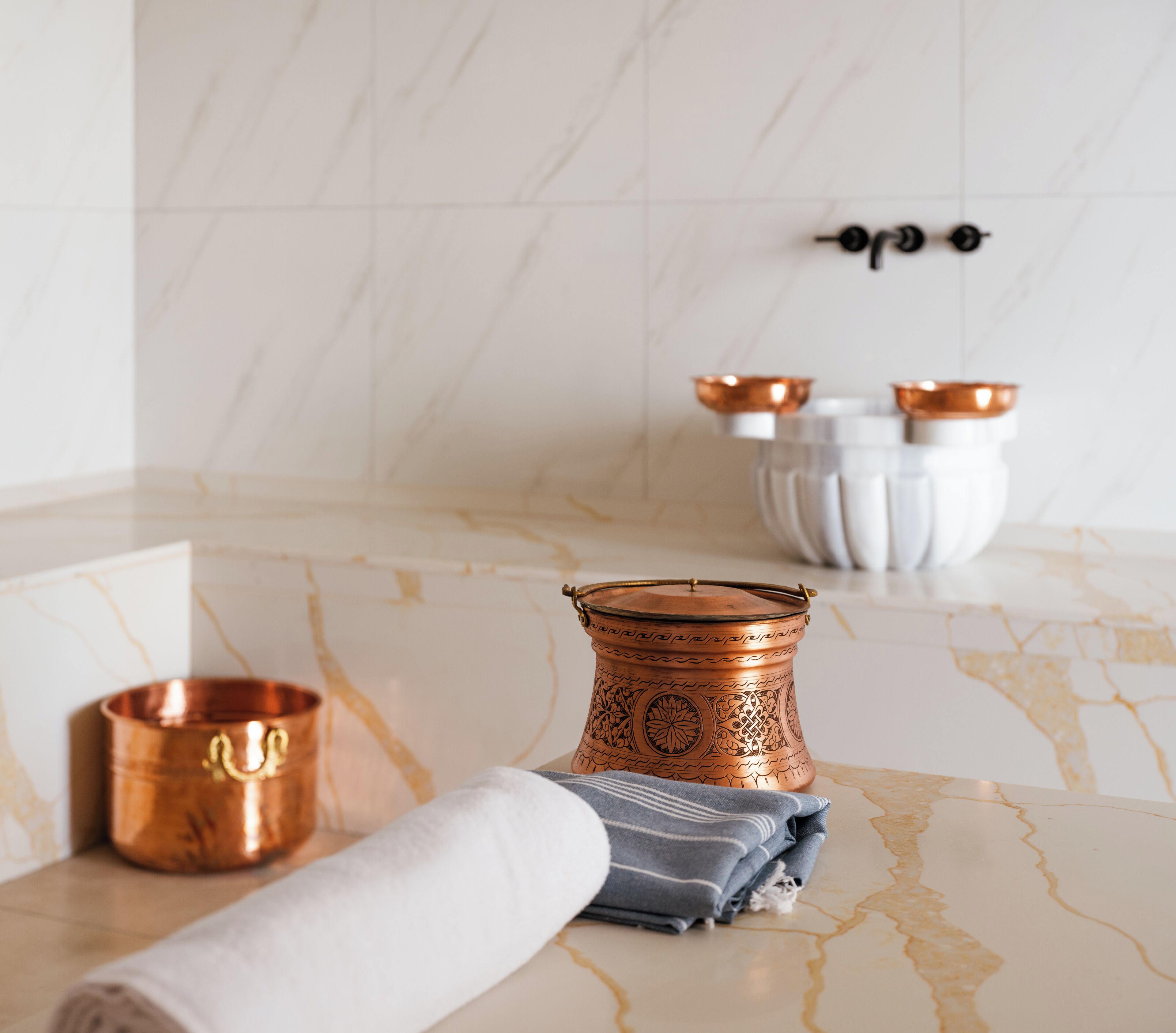 A bathroom with a marble floor, a copper vessel, a rolled towel, and a white basin with copper handles.