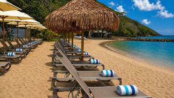 A beach with rows of sun loungers and thatched umbrellas, a clear blue sea, and a rocky coastline.