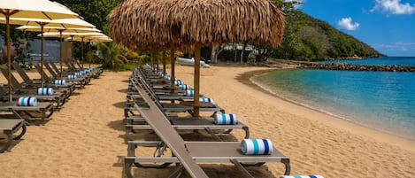 A beach with rows of sun loungers and thatched umbrellas, a clear blue sea, and a rocky coastline.