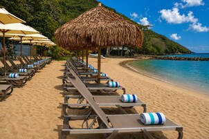 A beach with rows of sun loungers and thatched umbrellas, a clear blue sea, and a rocky coastline.