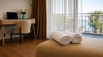 A hotel room with a wooden desk, a laptop, a chair, a bed with rolled towels, and a view of the sea through large windows.