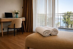 A hotel room with a wooden desk, a laptop, a chair, a bed with rolled towels, and a view of the sea through large windows.