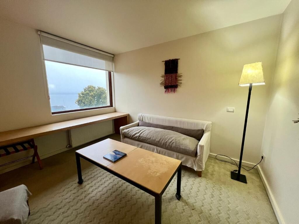A small, sparsely furnished room with a window, a sofa, a table, and a floor lamp.