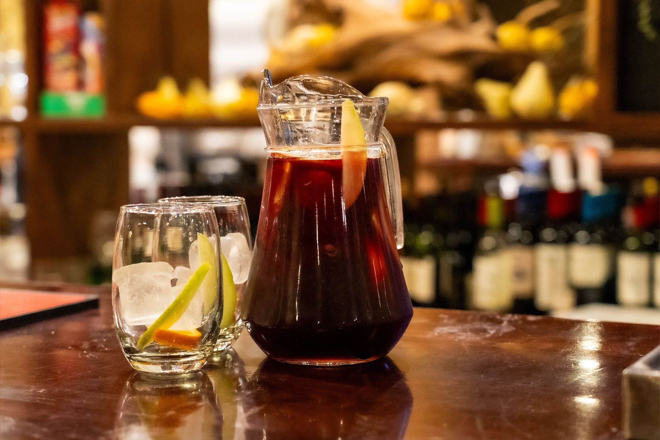 A bar setting with a pitcher of dark-colored beverage, two glasses with ice and citrus slices, and a blurred background with bottles.