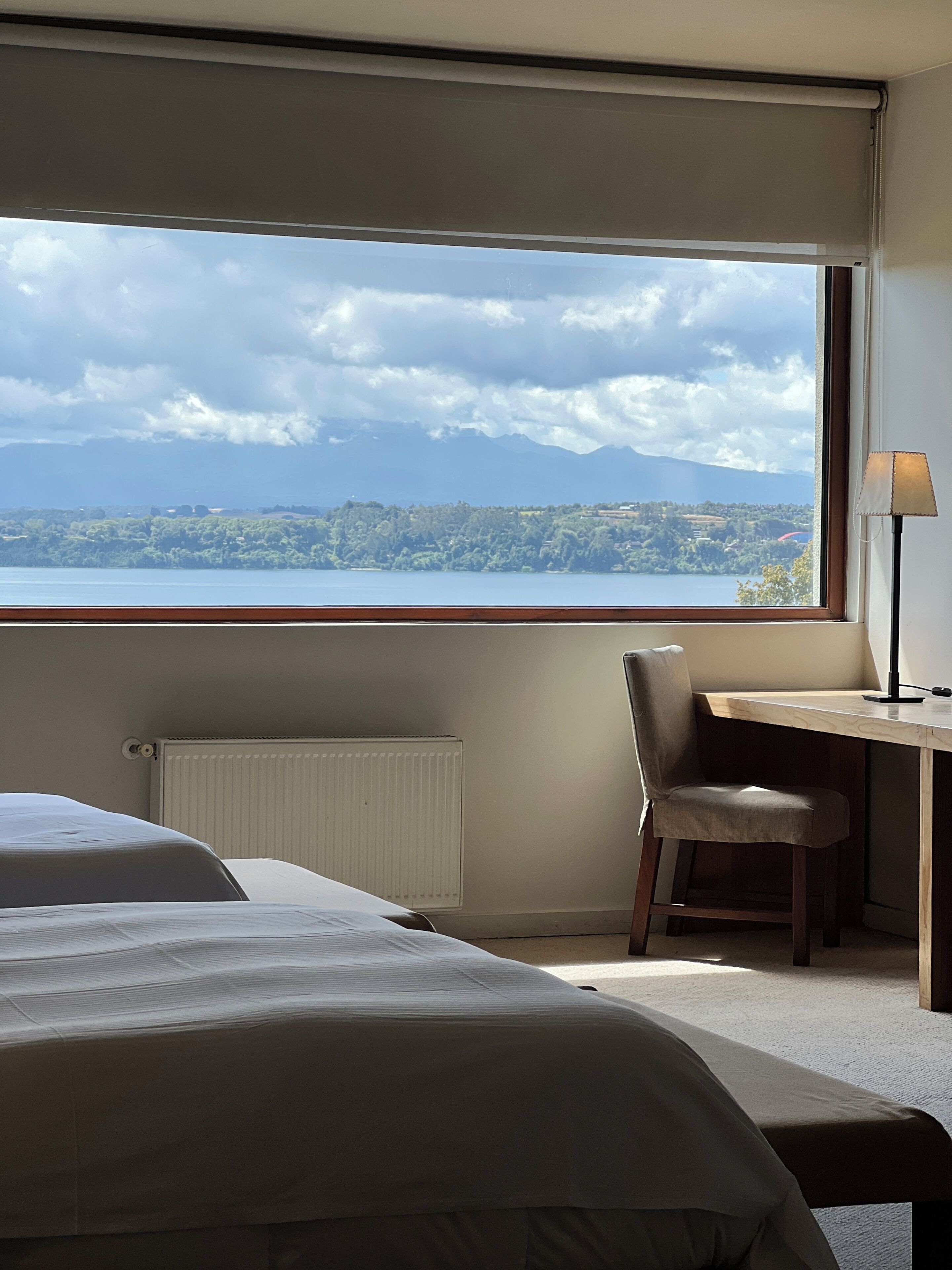 A hotel room with a large window offering a view of a lake and mountains, a desk with a chair, and a bed with a white duvet.