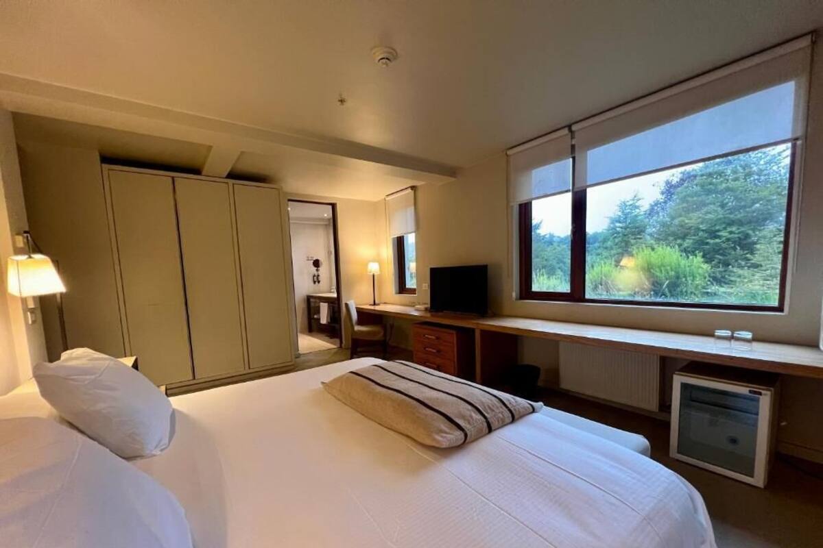 Ein Hotelzimmer mit Bett, Schreibtisch, Fernseher und einem Fenster mit Blick auf Grünflächen.