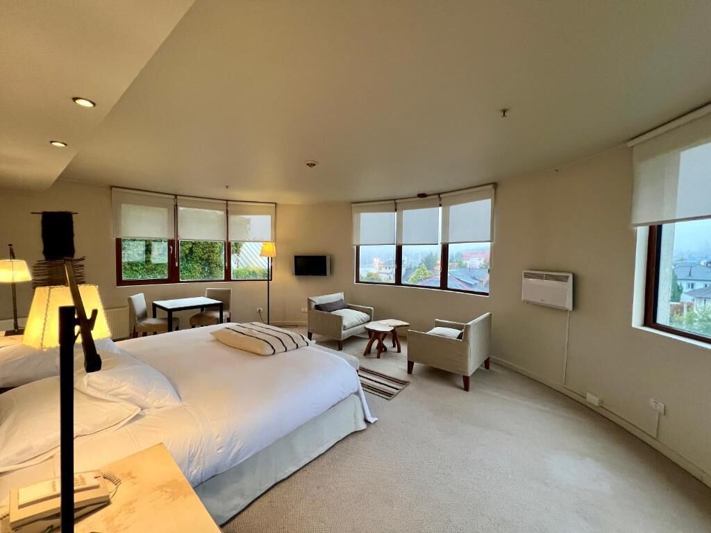 Habitación de hotel con una cama grande, un escritorio, dos sillas, un televisor y ventanas amplias.