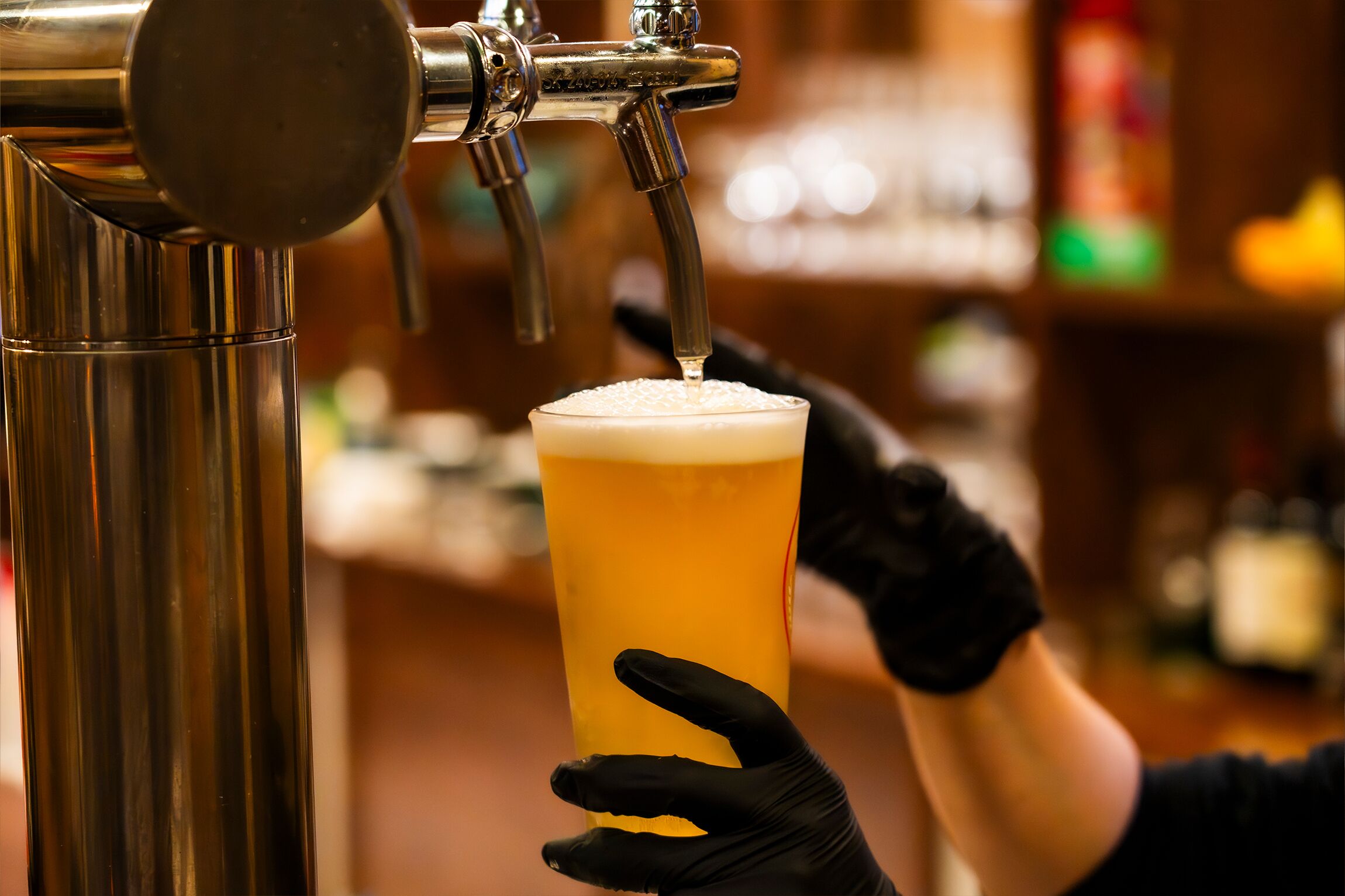 A tap dispensing beer into a glass held by a person wearing black gloves.