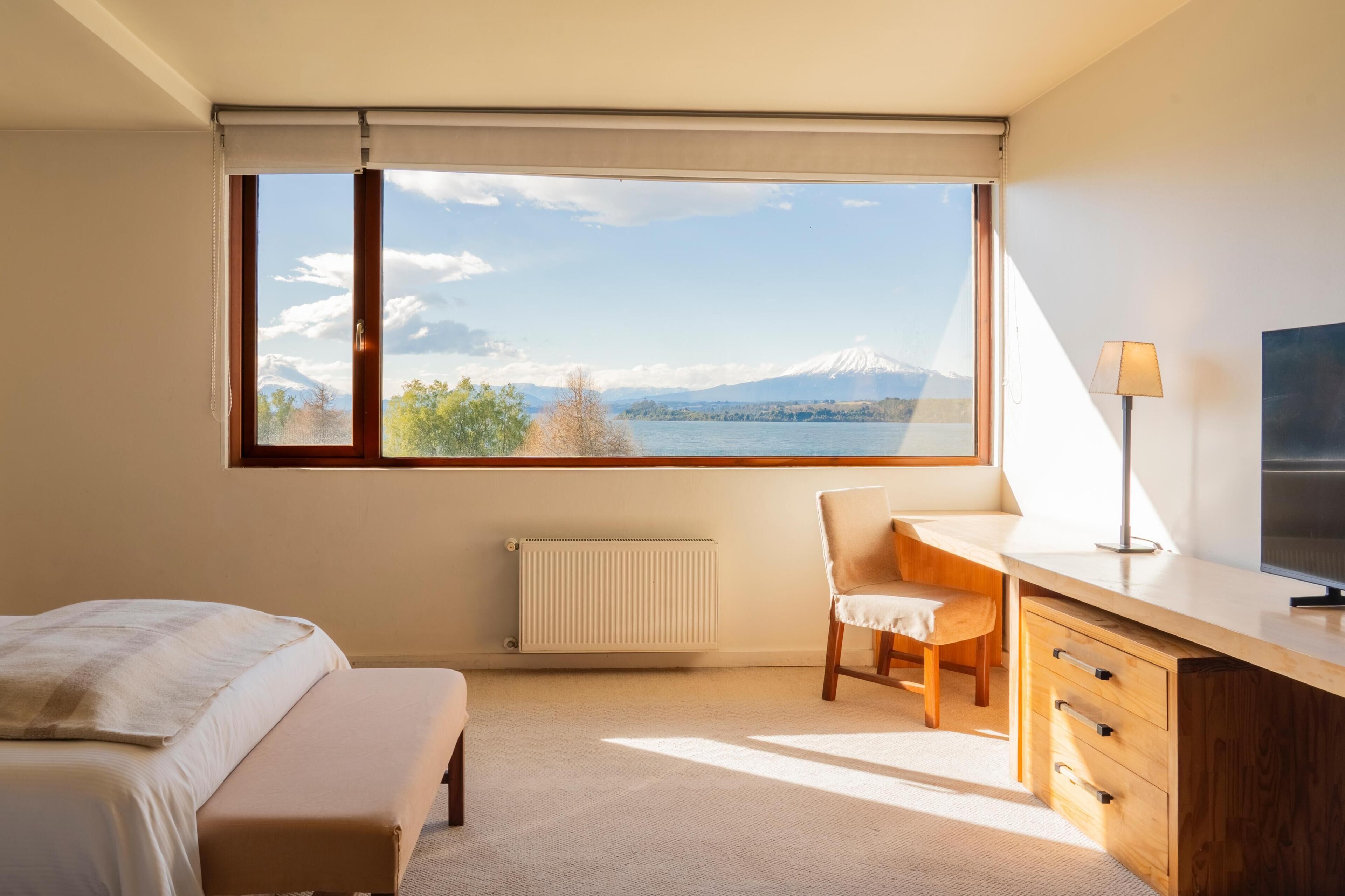 Ein Schlafzimmer mit Bett, Schreibtisch, Stuhl, Heizkörper und Blick auf einen Berg und See.