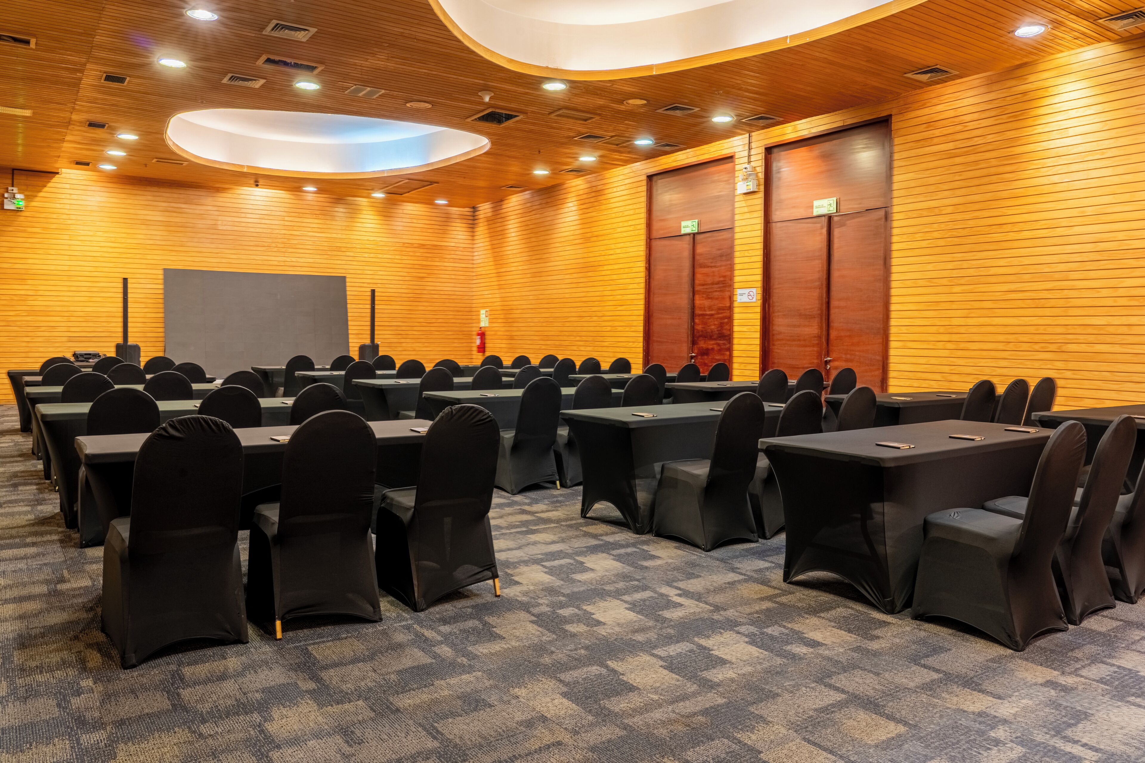 A conference room with rows of tables and chairs, wooden panel walls, and a ceiling with recessed lighting.