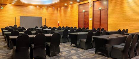 A conference room with rows of tables and chairs, wooden panel walls, and a ceiling with recessed lighting.