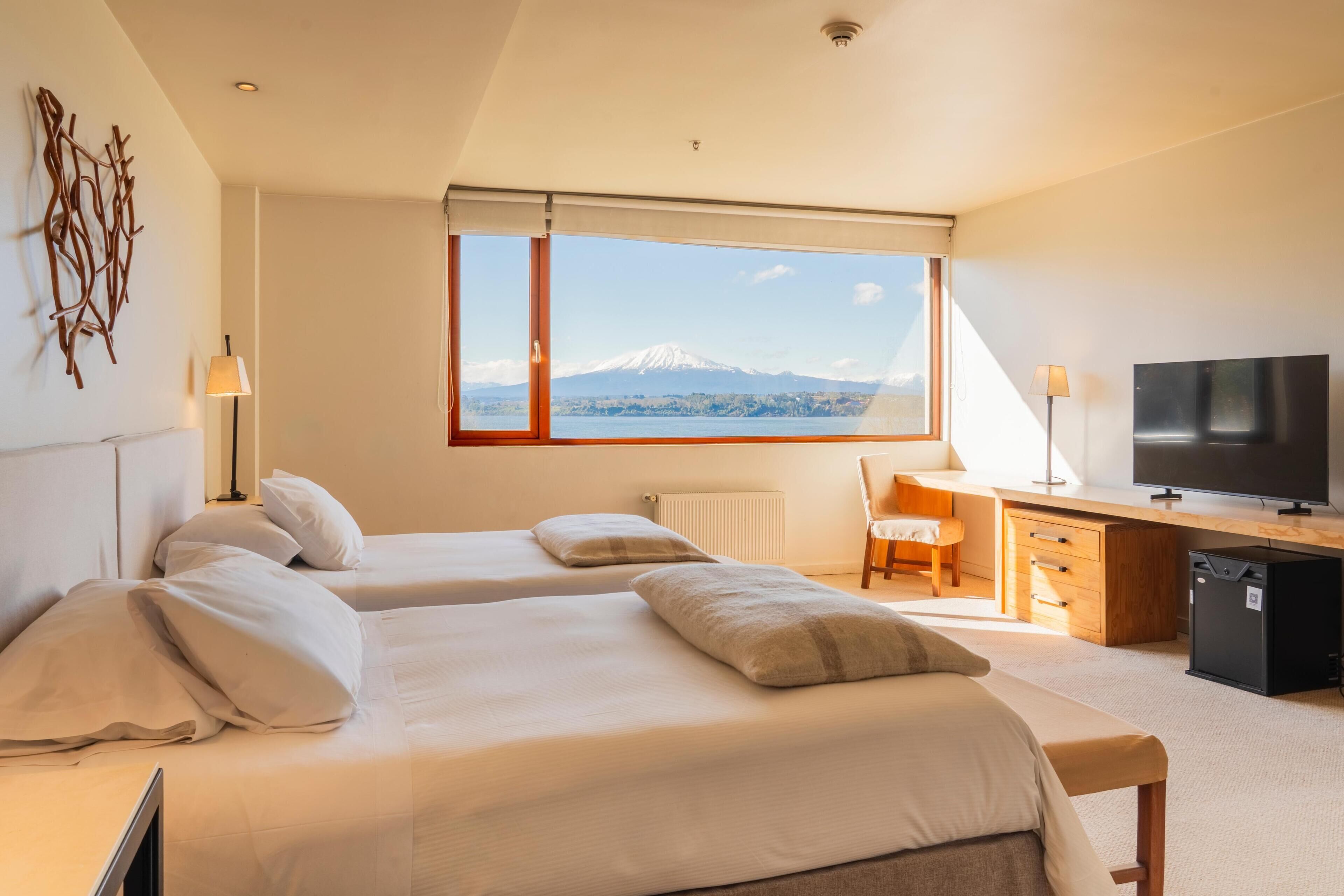 A hotel room with two beds, a desk, a chair, a TV, and a view of a mountain.