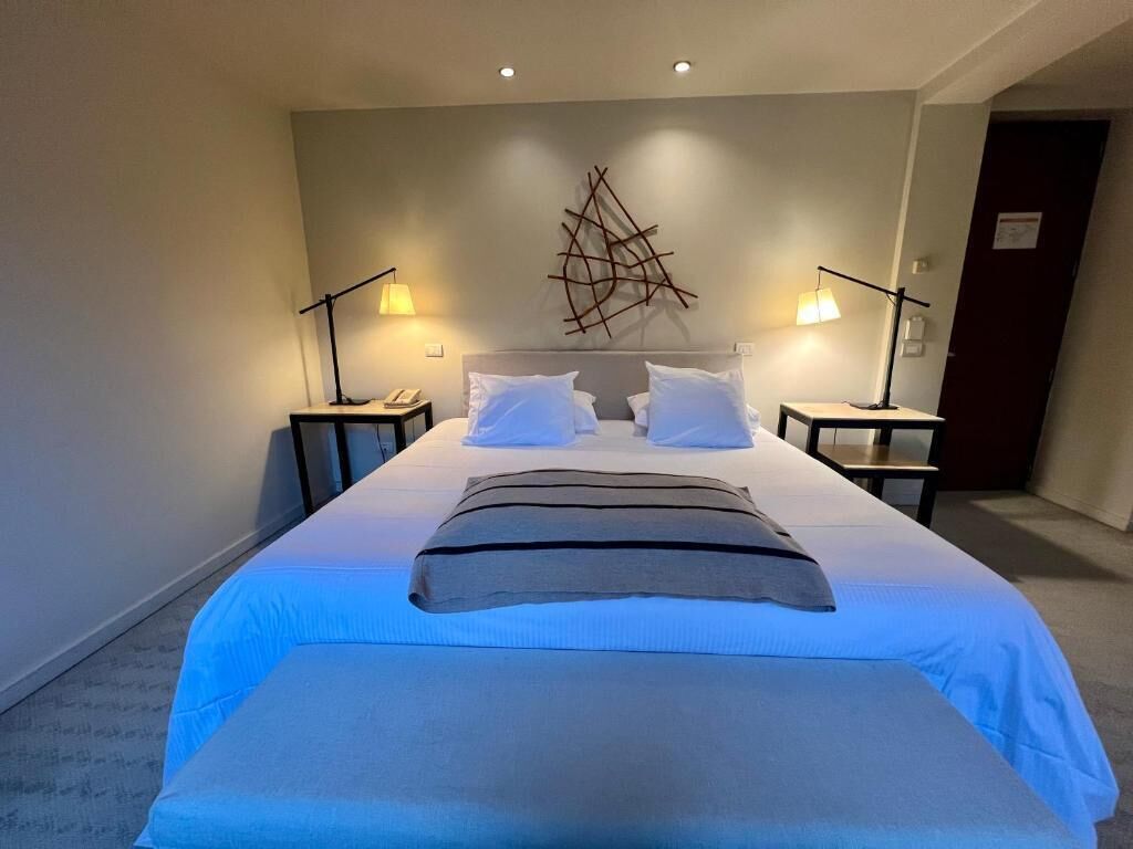 A hotel room with a large bed, two bedside tables with lamps, a wall-mounted abstract art piece, and a door in the background.