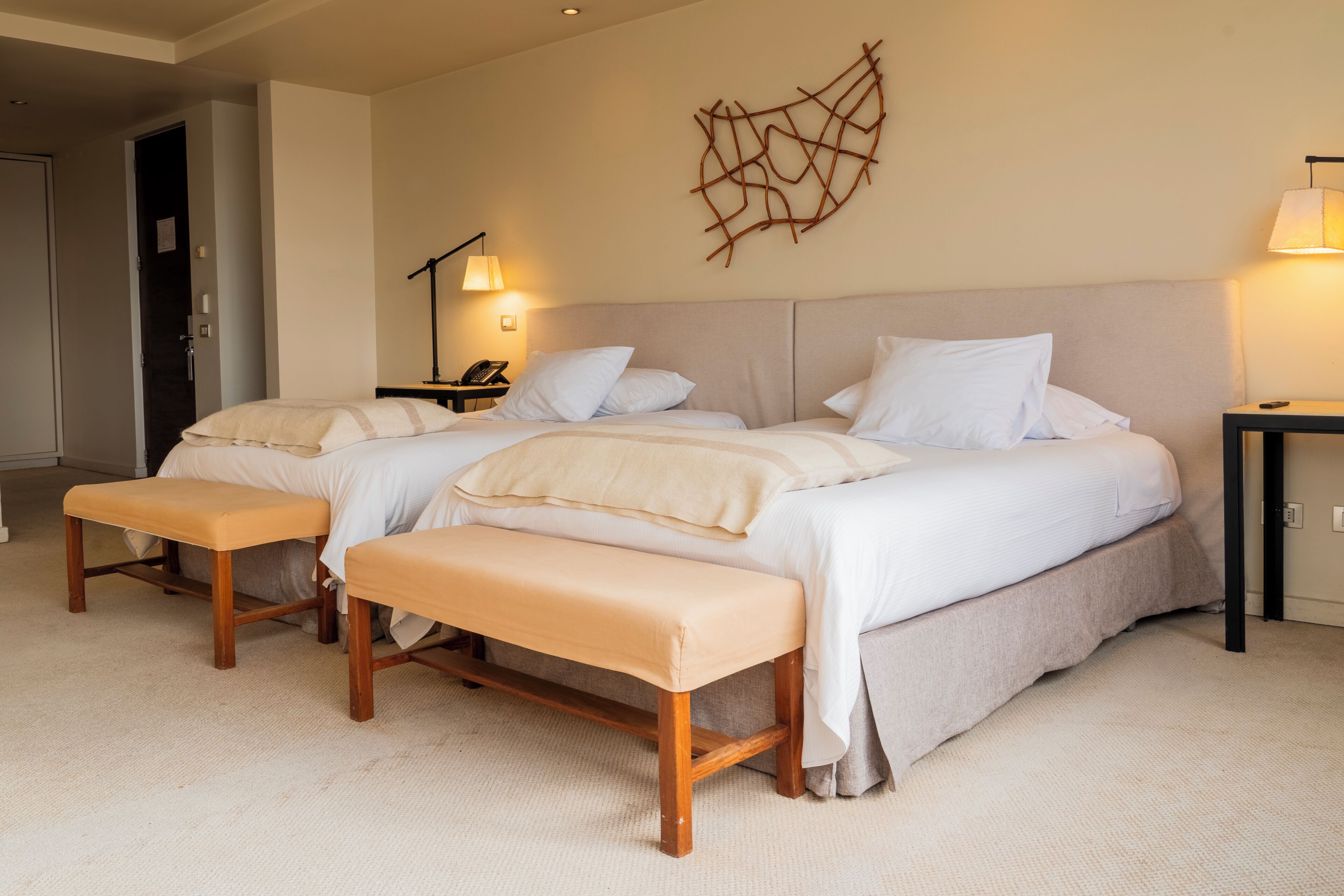 Hotelkamer met twee bedden, een bankje, een kunstwerk aan de muur, een nachtkastje met een lamp en een deur.