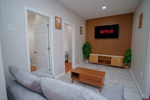 A living room with a sofa, coffee table, TV, wooden panel wall, and a door leading to another room. - NY Gateway | Arcade + Table Tennis | 15m to NYC (Jersey City)