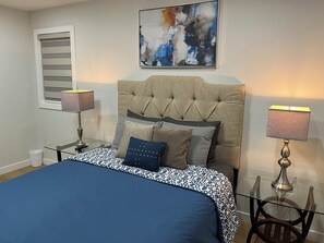 A bedroom with a bed, two bedside lamps, a glass table, and a painting on the wall.
