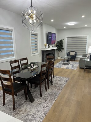 A modern dining area with a glass-top table, wooden chairs, a geometric pendant light, a fireplace, and a television on the wall.