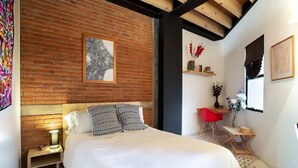 Ein Schlafzimmer mit Backsteinwand, einem Bett mit gemusterten Kissen, einem Nachttisch, einem roten Sessel, einem kleinen Tisch mit einer Vase und einem gerahmten Bild an der Wand.