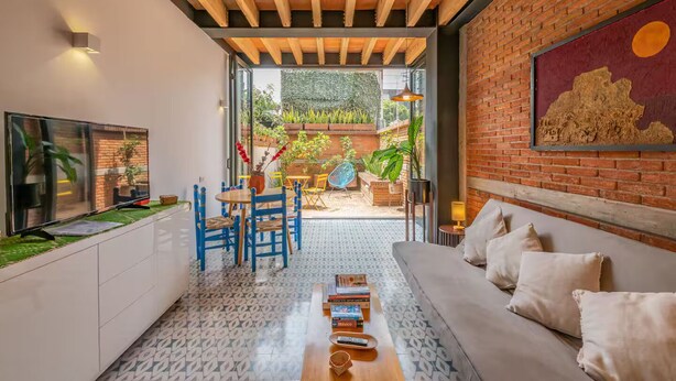 Ein modernes Wohnzimmer mit Sofa, Esstisch mit Stühlen, Fernseher und Bücherregal. Eine Ziegelwand ist mit einem Gemälde versehen, und durch eine offene Tür hat man Blick auf einen Garten.