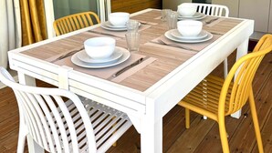 Comedor con una mesa dispuesta para cuatro, sillas blancas, una cocina con refrigerador y una puerta corrediza de vidrio.